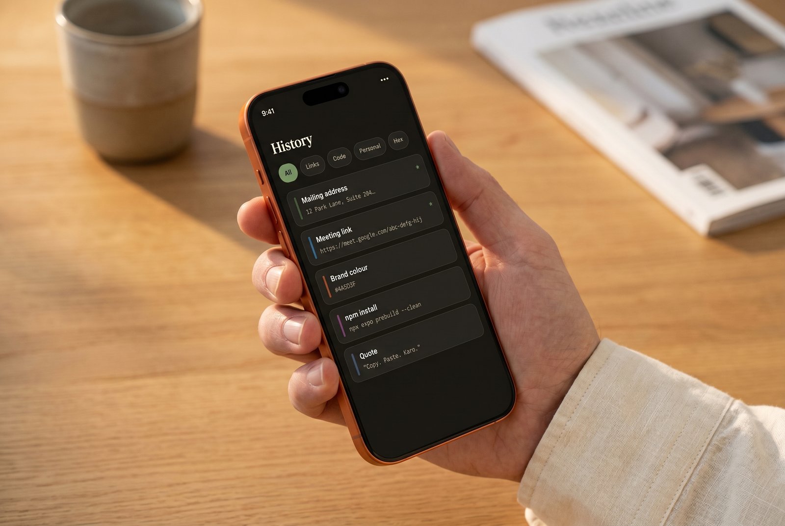 Copy-Paste Karo on iPhone 17 Pro — the History screen with a stack of saved snippets, held in one hand on a warm wooden desk.
